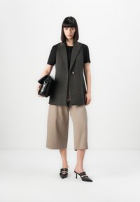 Vesta gri fără mâneci peste un tricou negru, pantaloni largi bej și ghete cu toc negre. Ținând o geantă de mână neagră. Design simplu.