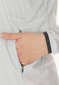 Lys grå jakke med et diskret prikmønster, lynlåslomme og elastiske manchetter. Materialet er glat og let.
