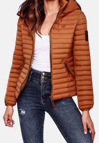 Hnědá prošívaná bunda s kapucí, vybavená zipovými kapsami a pružnými manžetami. Nositelná přes bílý top a v kombinaci s tmavými džínami.