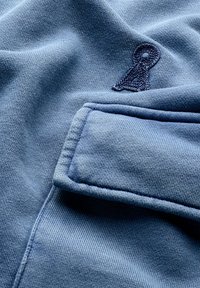 Blauer Baumwollstoff mit strukturiertem Oberflächen, einem kleinen gestickten Logo und einer genähten Taschenapplikation. Weiche und glatte Optik.