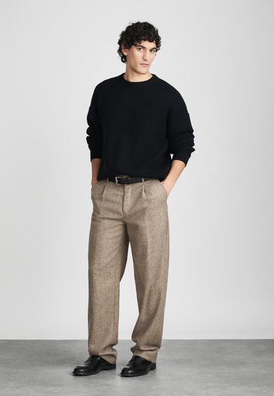 Zwarte ribgebreide trui, lichtbruine tweed broek met plooien, zwarte leren riem en zwarte dresschoenen. Eenvormige achtergrond.