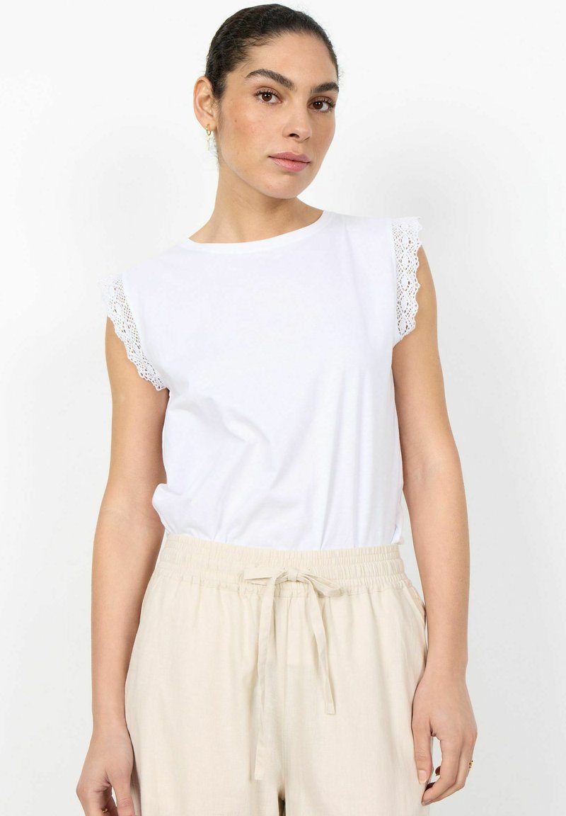 Femme aux cheveux foncés attachés, portant un haut blanc sans manches avec une bordure en dentelle et un pantalon beige à cordon, sur un fond blanc.