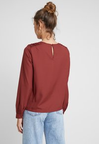 Bordeaux-paita, jossa on poimutus yksityiskohtina olkapäillä, pyöreä kaula-aukko, jossa on napitettu avaus, pitkät hihat ja sileä kangas.