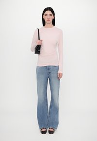 Light pink long-sleeve top with a fitted design, paired with loose blue jeans. Model holds a small black handbag and wears brown flats.