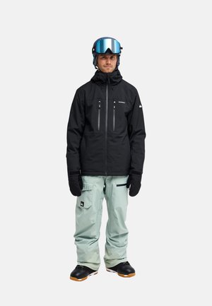 Mann trägt schwarze Skijacke, hellgraue Skihose, schwarze Handschuhe, schwarze Stiefel und blauen Skihelm mit reflektierenden Skibrillen, steht frontal.