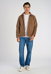 Brun kordflorjacka med dragkedja och två framfickor, bärs över en vitt t-shirt och blå jeans, med vita sneakers.