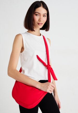 Mujer con el cabello lacio y oscuro, lleva un top blanco sin mangas y pantalones negros, y lleva un bolso bandolera rojo con un nudo en la correa.