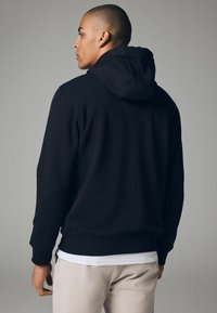 Sweat à capuche noir avec cordon de serrage, poignets et ourlet côtelés. Présente une texture lisse et une coupe décontractée, porté sur une chemise blanche et un pantalon beige.