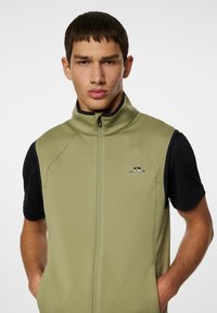 Olivgrön zip-up väst med hög krage, gjord av slätt tyg, med svarta kortärmar och en liten logotyp på bröstet.