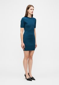 Γυναίκα στέκεται φορώντας ένα τιρκουάζ πλεκτό φόρεμα με κοντά μανίκια και υφή, μαζί με μαύρες μυτερές γόβες, μπροστά σε λευκό, μονόχρωμο φόντο.