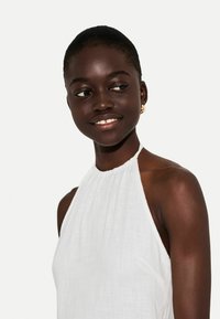 Vit halterneck-klänning i texturerat tyg, med en samlad halsringning och öppen rygg, bärs med guldörhängen i hoop-stil.