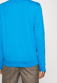 Sudadera azul con mangas largas y cuello acanalado. Cuenta con una cintura texturizada con la marca "LACOSTE". El material parece suave y liso.