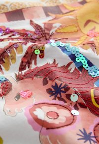 Tissu coloré avec broderie complexe et paillettes représentant des animaux stylisés et des motifs floraux dans des tons chauds.