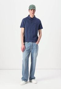 Polo azul-marinho com um padrão texturizado, combinado com calças de ganga largas de um azul claro e sapatilhas brancas. Os acessórios incluem um chapéu verde.