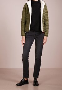 Grøn pufferjakke med hvid fleece-fôr, sort sweater, slanke sorte jeans og sorte sko. Jakken har lynlås og elastiske manchetter.