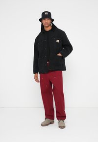 Čierna denimová bunda s vreckami a logom Carhartt, skombinovaná s tmavočervenými nohavicami a šedými čižmami, doplnená čiernym vedrom.