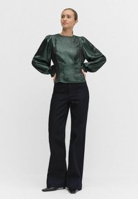 Zielona, błyszcząca bluzka z bufiastymi rękawami, w zestawieniu z ciemnymi, szerokimi spodniami. Bluzka ma okrągły dekolt i elastyczne mankiety.
