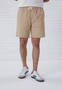 Costumini da bagno beige con vita elasticizzata e cordino bianco. Indossati con sneaker bianche con accenti neri e suole grigie.