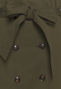 Oliwkowy płaszcz trench z podwójnym zapięciem, dużymi brązowymi guzikami oraz przewiązywanym paskiem. Gładka struktura materiału i dopasowany krój.