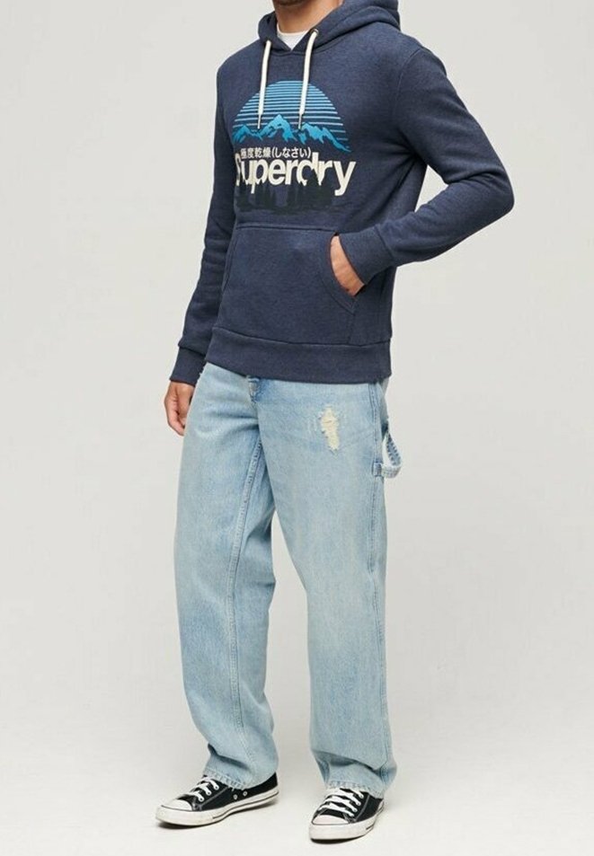 Homme portant un sweat à capuche Superdry bleu marine avec un motif de montagne, un jean bleu clair déchiré et des baskets basses noires à semelles blanches.