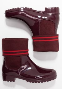 Botas de lluvia de tobillo en un brillante burdeos oscuro con puños de punto que presentan dos franjas rojas horizontales, mostradas desde un lado y vista inferior sobre fondo blanco.