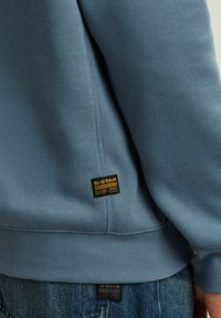 Felpa azzurra chiara con polsini e orlo a costine, decorata da una toppa con logo nero e giallo sul lato sinistro. Il tessuto sembra morbido.