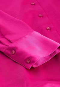 Fuchsia Langarmhemd mit glatter Textur, ausgestattet mit zwei geknöpften Manschetten und goldfarbenen Knopfakzenten entlang der vorderen Patte.