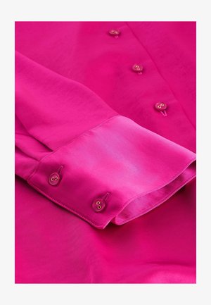 Chemise fuchsia à manches longues avec une texture lisse, dotée de deux poignets à boutons et d'accents de boutons dorés le long de la patte de boutonnage.