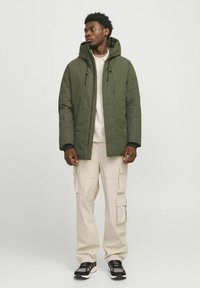 Abrigo verde con capucha y cierre de cremallera, combinado con pantalones cargo en color beige claro. Zapatillas con detalles en negro y blanco. Diseño y ajuste minimalista.