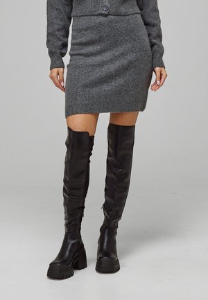 Femme portant une mini-jupe en maille grise et un cardigan assorti avec des bottes noires à semelles épaisses et plateforme jusqu'aux genoux sur un fond uni.