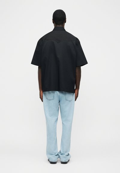 Muž stojící zády, oblečený v volné černé košili s krátkým rukávem a světle modrých džínech s rovnými nohavicemi, na jednobarevném pozadí.