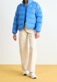 Blå puffjacka med dragkedja fram, elastiska ärmmuddar och logodetalj. Bärs över en blå topp med beige vida byxor och vita sneakers.