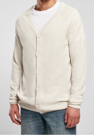 Man draagt een off-white gestructureerde gebreide vest met knopen over een wit t-shirt en blauwe spijkerbroek, afgebeeld van nek tot heupen.