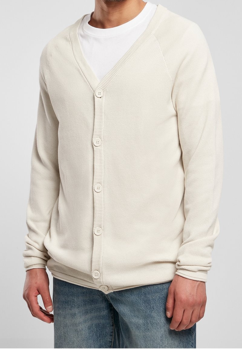 Homme portant un cardigan texturé blanc cassé boutonné sur un t-shirt blanc et un jean bleu, visible du cou jusqu'aux hanches.