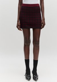 Gebreide mini rok in bordeauxrood en zwart, met een gestructureerd ruitpatroon, elastische tailleband en een aansluitende pasvorm. Gecombineerd met zwarte sokken en puntige schoenen.