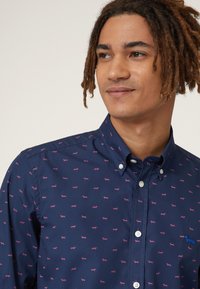 Marineblauwe overhemels met kleine roze hondenpatronen, voorzien van een kraag en een blauw geborduurd hondenlogo op de borst. Gladde stoftextuur.