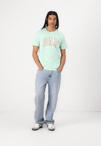 Camiseta verde menta con el logo "GAP" en naranja, combinada con jeans rectos de un azul claro y zapatillas casuales grises y blancas.
