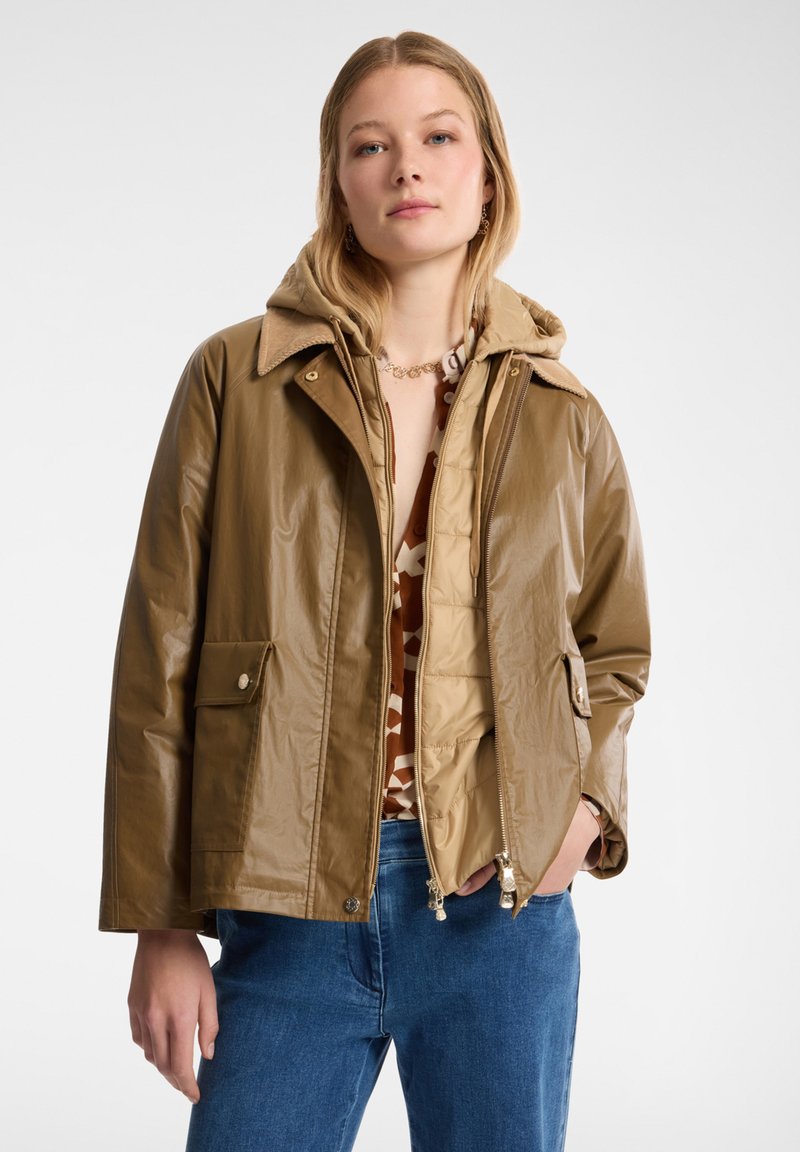 Giacca impermeabile marrone con cappuccio, chiusura con zip, due tasche frontali e un rivestimento interno trapuntato. Presenta una texture liscia e lucida.