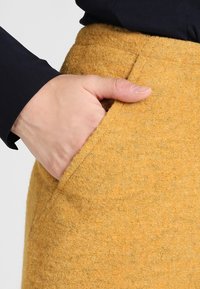 Jupe en laine couleur jaune moutarde, avec une poche latérale et une finition douce et texturée. Main placée dans la poche.
