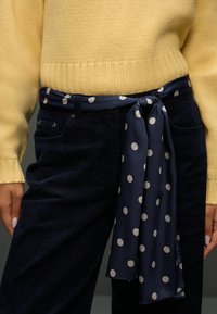 Granatowy szalik w groszki zawiązany w pasie, zestawiony z żółtym, dzianym swetrem i ciemnymi sztruksowymi spodniami. Zbliżenie na tekstury tkanin.