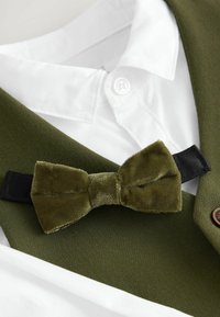 Noeud papillon de couleur vert olive en velours texturé, fixé à une bande sombre. Positionné sur une chemise col blanc et un gilet en tissu vert.