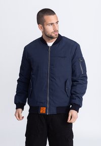 Marineblauwe bomberjack met een ritssluiting aan de voorkant, geribbelde boorden, zijzakken en een oranje label aan de zoom. Soepele textuur, lichtgewicht materiaal.