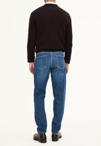 Dunkelbrauner Pullover, blaue Jeans, schwarze Schuhe. Der Pullover hat einen runden Ausschnitt und gerippte Bündchen. Die Jeans haben einen geraden Schnitt mit Gesäßtaschen.