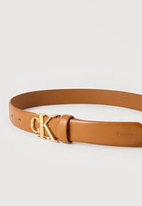 Ζώνη από δέρμα καμήλας με χρυσό αγκράφα "cK" και ανάγλυφο λογότυπο Calvin Klein κοντά στο τέλος, τοποθετημένη επίπεδα σε λευκή επιφάνεια.