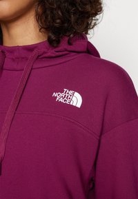 Gros plan d'une personne portant un sweat à capuche violet avec des cordons et un logo blanc The North Face sur la poitrine.