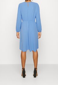 Himmelblaues, knielanges Kleid mit langen Ärmeln, elastischem Taillenbund und einem Schlüsselloch-Detail im Rücken. Kombiniert mit schwarzen, hochhackigen Schuhen.