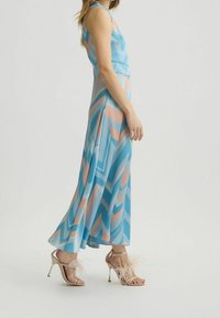 Robe maxi bleue avec des motifs en chevron rose et blanc, fabriquée à partir d'un tissu lisse, associée à des talons dorés à lanières ornés de plumes.