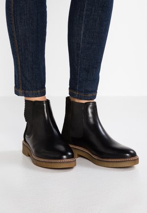 Person trägt dunkelblaue Skinny-Jeans und schwarze Leder-Stiefeletten mit Gummisohlen und steht auf einer weißen Fläche.