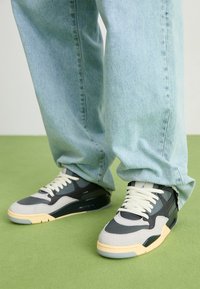 Grå og sorte sportssko med lysegrå detaljer og cremefarvede snørebånd. Båret med løse lys vaskede denimjeans. Grøn baggrundsflade.