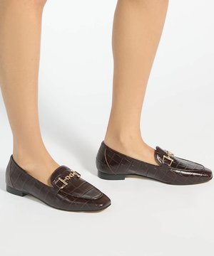 Pieds portant des mocassins en cuir embossé croco marron foncé avec détail chaîne dorée sur fond blanc.